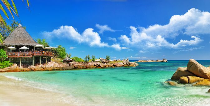 Strand mit Sand und Felsen, links ein Restaurant mit Strohdach und Sonnenschirmen, türkisfarbenes Meer und blauer Himmel mit Wolken