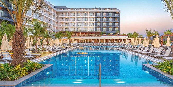 Großer rechteckiger Hotelpool mit Liegestühlen und Sonnenschirmen an den Seiten vor einem mehrstöckigen Hotelgebäude bei Sonnenuntergang