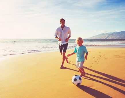 Vater und Sohn spielen barfuß am Strand Fußball bei klarem Himmel und beginnendem Sonnenuntergang.