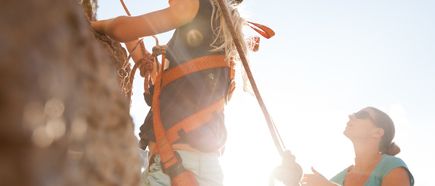 Kind mit Kletterausrüstung klettert an einer Felswand während eine erwachsene Person sichert