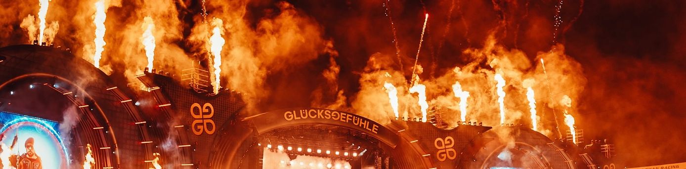 Bühne eines Festivals mit Feuerwerk und Flammen, davor große Menschenmenge, Schriftzug 'GLÜCKSGEFÜHLE' sichtbar
