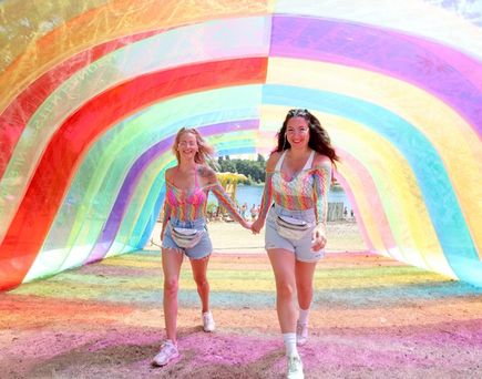 Zwei Frauen in kurzen Hosen und bunten Oberteilen laufen Hand in Hand durch einen Tunnel aus Regenbogenfarben auf einem Festivalgelände
