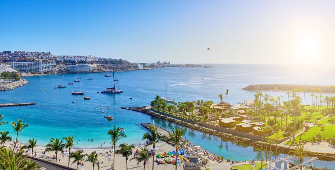 Blick auf eine Küstenlandschaft mit Sandstrand, Palmen, Booten im Wasser und einem Sonnenuntergang im Hintergrund.