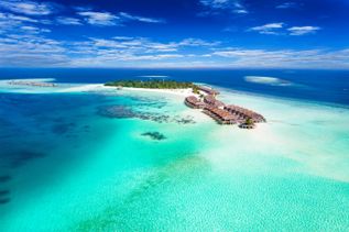 Luftaufnahme einer tropischen Insel mit Sandstrand, umgeben von türkisfarbenem Wasser und mehreren über dem Wasser gebauten Bungalows