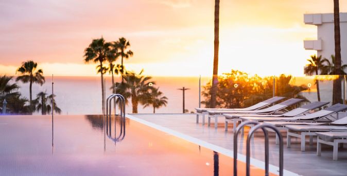Ein Pool mit einem reflektierenden Wasseroberfläche im Vordergrund, umgeben von Liegen und Palmen, während die Sonne am Horizont untergeht.