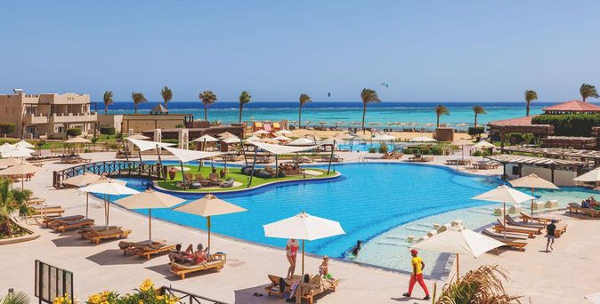 Großer Hotelpool mit Liegen und Sonnenschirmen, umgeben von Gebäuden und Palmen, im Hintergrund das Meer unter klarem Himmel