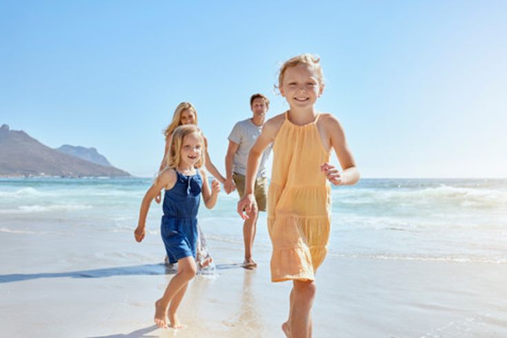 Familie mit zwei Kindern läuft barfuß lachend am Strand entlang, im Hintergrund Meer und Berge