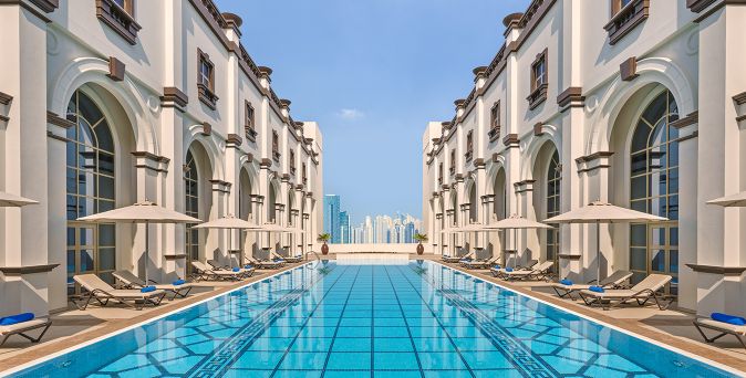 Symmetrischer Außenpool zwischen zwei mehrstöckigen Gebäudeflügeln mit Liegestühlen und Blick auf die Skyline von Dubai