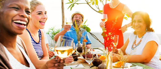 Gruppe von fünf Personen sitzt an gedecktem Tisch im Freien, hebt Weingläser zum Anstoßen, Tisch mit Blumen und Snacks dekoriert