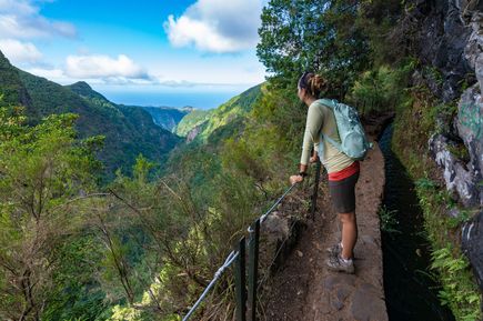 Person auf dem Wanderweg Levada do Alecrim auf Madeira mit Blick über ein grünes Tal, bewaldete Berghänge und den Atlantik in der Ferne