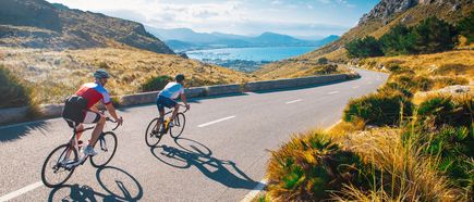 Zwei Radfahrer auf einer kurvigen Bergstraße mit Blick auf das Meer und die Bucht, unter blauem Himmel