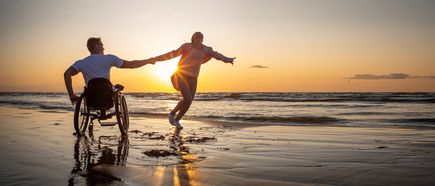 Zwei Personen bewegen sich gemeinsam am Strand bei Sonnenuntergang, einer davon sitzt im Rollstuhl, während die andere die Hand hält