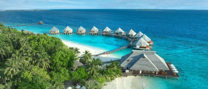 Bungalows auf Stelzen über türkisfarbenem Wasser, verbunden durch Stege, umgeben von dichtem Grün und weißem Sandstrand am Adaaran Select Medhupparu Resort.