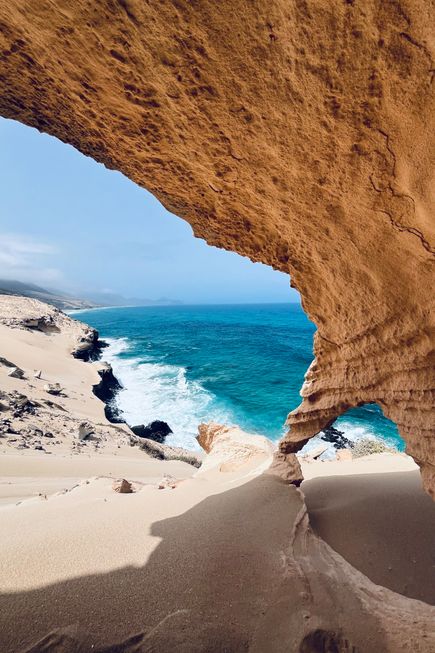Felsformation mit natürlichem Bogen am Strand von Fuerteventura, dahinter Meer und bewölkter Himmel.