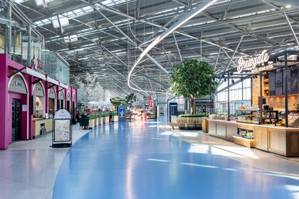 Moderne Innenansicht des Terminal 1 am Flughafen Köln/Bonn mit breitem Passagierbereich, Gastronomie- und Einkaufsmöglichkeiten, markanten Lichtinstallationen, Sitzgelegenheiten und großzügiger Glas-Stahl-Architektur für Reisende vor dem Abflug.