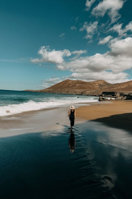 Frau mit Hut und schwarzem Kleid geht am Strand von Fuerteventura entlang, Berge im Hintergrund, bewölkter Himmel.