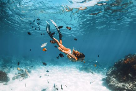 Frau beim Schnorcheln unter Wasser, umgeben von bunten tropischen Fischen und Korallen in klarem, türkisblauem Meer