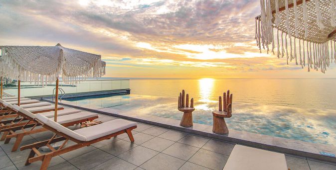 Infinity-Pool mit zwei großen Holzhand-Skulpturen am Beckenrand und Liegestühlen unter Sonnenschirmen bei Sonnenuntergang.