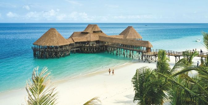 Steg des Hotel Riu Jambo auf Sansibar mit mehreren strohgedeckten Pavillons über türkisfarbenem Wasser, weißer Sandstrand und zwei Personen am Ufer