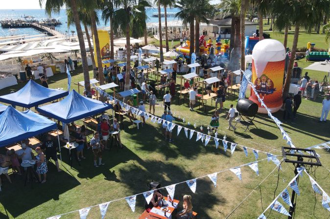 Oktoberfest-Veranstaltung in einer weitläufigen Hotelanlage am Strand mit festlich dekorierten Biergartenständen, zahlreichen Gästen, Palmen und einer aufblasbaren Bierkrug-Figur mit der Aufschrift „Otto Beer Fest“.