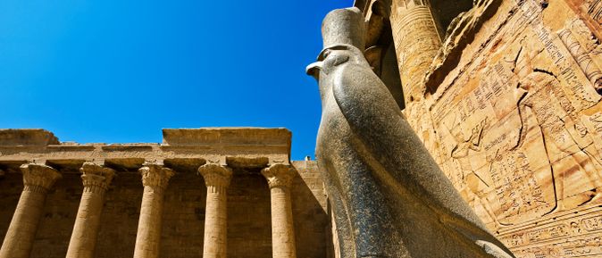 Große steinerne Statue des Gottes Horus mit Falkenkopf vor Säulengang und Wand mit Hieroglyphen im Tempel von Edfu