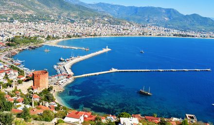 Luftaufnahme des Hafens von Alanya in der Türkei mit geschwungenem Wellenbrecher, mehreren Booten im türkisblauen Meer