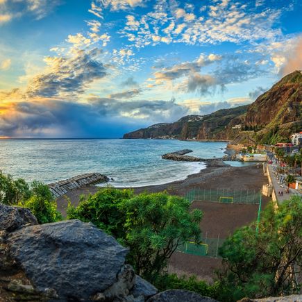 Küstenort Ribeira Brava auf Madeira bei Sonnenuntergang mit dunklem Kiesstrand, Wellenbrechern, Steilküste und Blick über den Atlantik unter einem wolkenreichen Himmel
