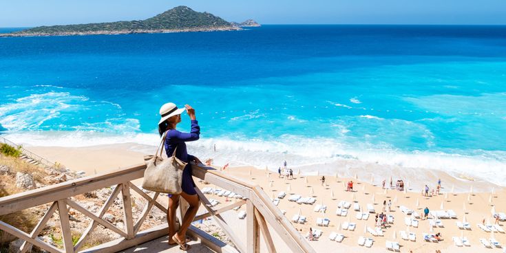 „Frau mit Strohhut blickt auf den Kaputas-Strand in der Türkei mit türkisblauem Meer, Sandstrand und Sonnenliegen