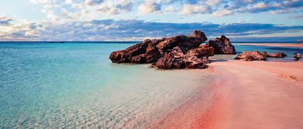 Klares türkisfarbenes Meer mit flachem Sandstrand und einer Gruppe brauner Felsen unter bewölktem Himmel.