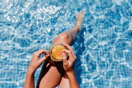 Frau sitzt entspannt am Poolrand und hält ein Getränk über dem klaren Wasser eines gefliesten Hotelpools