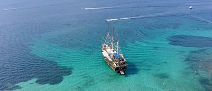 Piratenähnliches Segelschiff mit zwei Masten auf türkisblauem, klarem Wasser, umgeben von dunklen Schatten unter Wasser.