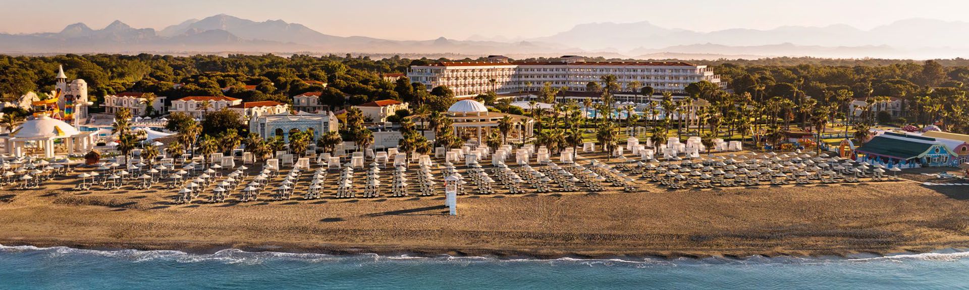 Strand mit Liegestuhlreihen und Sonnenschirmen vor dem Hotel Selectum Noa Belek, dahinter Pinienwald und Berge im Hintergrund.