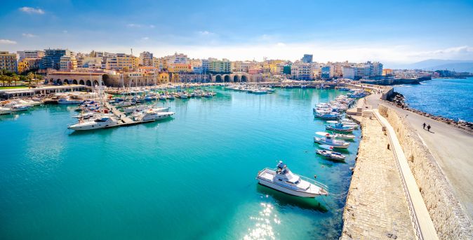Hafen von Heraklion auf Kreta mit mehreren Booten im Wasser und einer Ufermauer, im Hintergrund Stadtgebäude unter klarem Himmel.