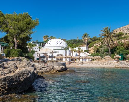 Küstenbereich mit Felsen, klarem Wasser, weißen Gebäuden mit Kuppeldach und mehreren Sonnenschirmen, umgeben von Palmen und Pinien auf Rhodos.