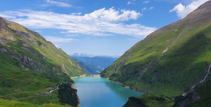 Blick auf den türkisfarbenen Stausee in der Region Kaprun im Salzburger Land, umgeben von grünen Bergen unter blauem Himmel mit Wolken.