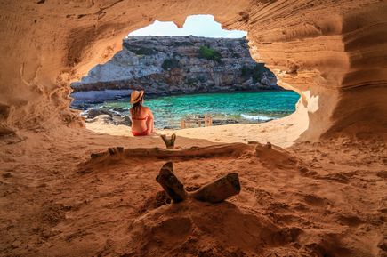 Frau in rotem Bikini und Strohhut sitzt am Eingang einer Felsenhöhle und blickt auf die türkisfarbene Bucht Cala en Baster auf Formentera