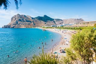Strand mit Badegästen und Hotels vor felsigem Gebirge an der Küste von Kolymbia auf Rhodos.
