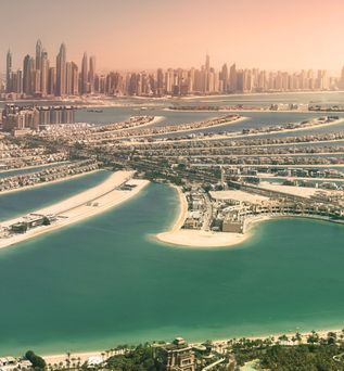 Skyline der Vereinigten Arabischen Emirate. Im Vordergrund das Meer und im Hintergrund eine Landzunge mit mehreren Wolkenkratzern. Der Himmel ist leicht rosa gefärbt.