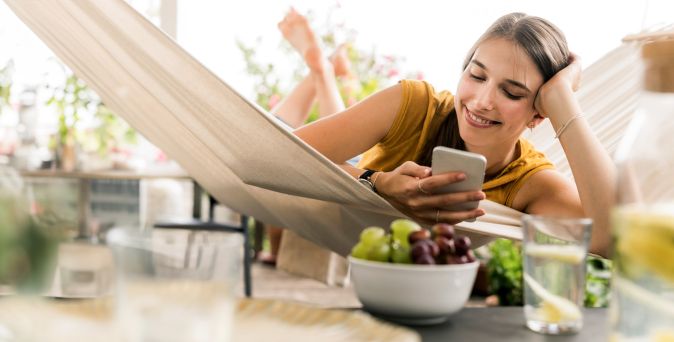 Frau liegt lächelnd in einer Hängematte und hält ihr Smartphone in der Hand