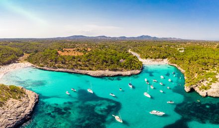Blick auf die Bucht Cala Mondragó auf Mallorca mit türkisfarbenem Wasser, bewaldeten Küsten, Sandstrand und zahlreichen Booten in einer naturbelassenen Umgebung