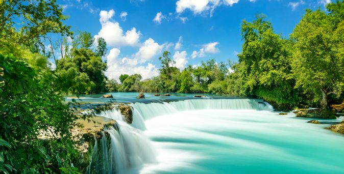 Breiter Wasserfall mit türkisfarbenem Wasser umgeben von dichtem grünen Wald unter blauem Himmel mit weißen Wolken
