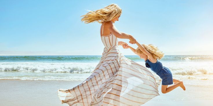 Frau in einem langen Sommerkleid schwingt ein Kind am Strand, während im Hintergrund Wellen an den Sand rollen