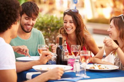 Mehrere Personen sitzen gemeinsam an einem gedeckten Tisch im Freien, genießen Essen und Getränke und unterhalten sich in entspannter Atmosphäre
