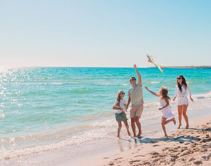 Vier Personen am Strand, zwei Kinder und zwei Erwachsene, spielen mit einem bunten Drachen am Meer bei klarem Himmel.