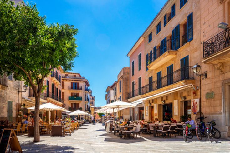 Belebte Gasse in der Altstadt von Alcúdia auf Mallorca mit Straßencafés, Sonnenschirmen und historischen Gebäuden