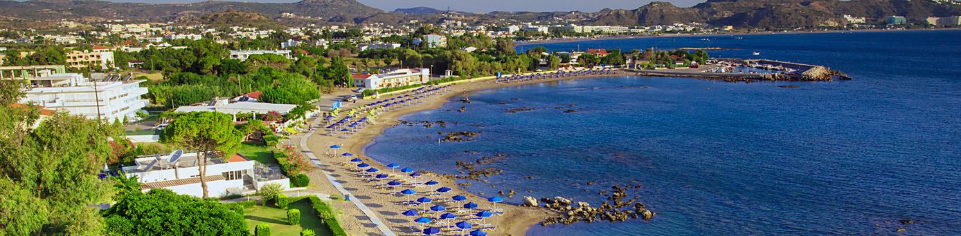Luftaufnahme eines gebogenen Sandstrands mit mehreren blauen Sonnenschirmen und Liegen, umgeben von Gebäuden und Bergen im Hintergrund.