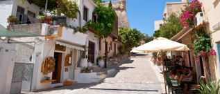 Schmale Gasse in Rethymnon auf Kreta mit weiß getünchten Häusern, blühenden Bougainvillea und Menschen unter einem Sonnenschirm an einem Straßencafé.