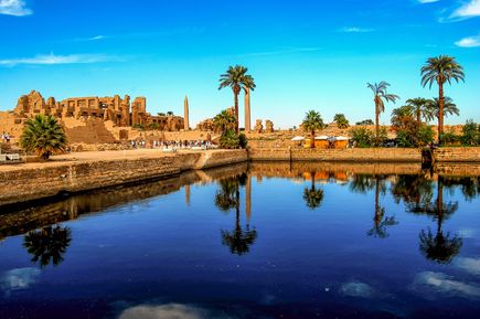 Blick auf den Karnak-Tempel in Luxor, Ägypten, mit antiken Ruinen, Palmen und einem Wasserbecken, das die Tempelanlage und den blauen Himmel spiegelt