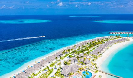 Resort auf den Malediven mit Wasservillen über einer türkisblauen Lagune, einem langen Sandstrand, Palmen und tiefblauem Ozean im Hintergrund