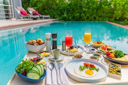 Schwimmendes Frühstück mit Eierspeise, frischem Obst, Brot, Salat und Säften auf einem Tablett in einem Pool vor einer Terrasse mit Liegen